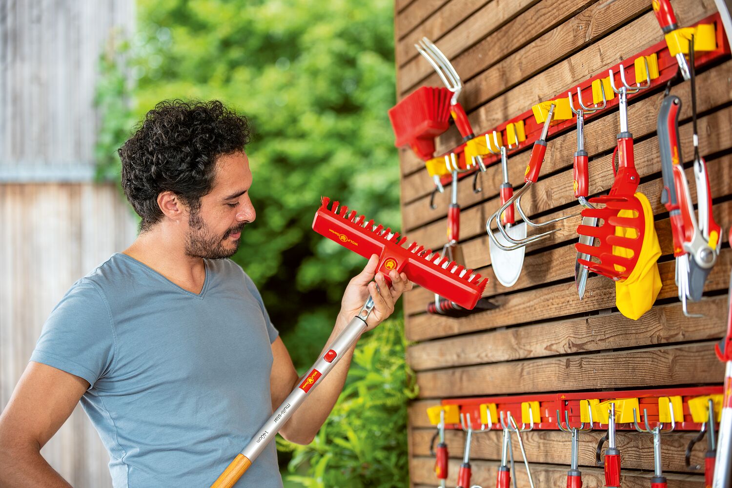 Man holding a garden tool next to a wall-mounted tool rack with various gardening tools.